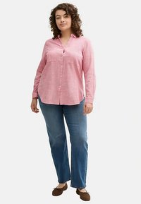 Camicia a maniche lunghe con bottoni in rosa chiaro, realizzata in tessuto strutturato con finitura morbida e due tasche sul petto, abbinata a jeans blu e scarpe marroni.