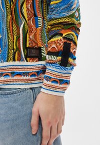 Värikäs neulepaita, jossa on monimutkaisia kuvioita sinisissä, oransseissa ja vihreissä sävyissä, ribattu hihansuut ja musta brändilabel yksityiskohta.