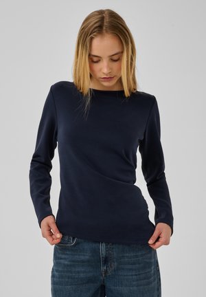 Jeune femme aux cheveux blonds raides portant un haut sombre uni à manches longues et un jean bleu, qui regarde vers le bas en tenant l'ourlet du haut.