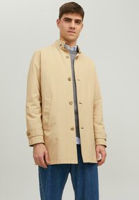 Beige, lichtgewicht trenchcoat met een staande kraag, voorknopsluiting en zijzakken, gedragen over een grijze trui en blauwe jeans.