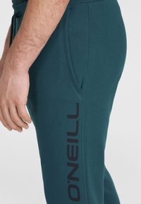 Skrócone dresy w kolorze teal, wykonane z miękkiego materiału, o zwężanym kroju, z boczną kieszenią i dużym czarnym logo nadrukowanym wzdłuż uda.