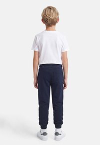 Ξανθός αγόρι που βλέπει προς τα πίσω, φοράει λευκό κοντομάνικο πουκάμισο, ναυτικό τζόγκερ παντελόνι με πίσω τσέπη, λευκές κάλτσες και λευκά αθλητικά παπούτσια.