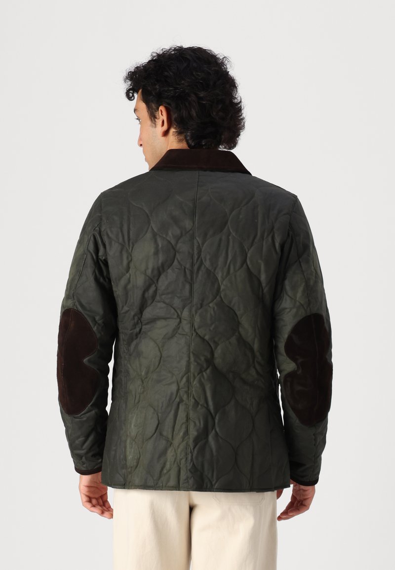 Grüne Steppjacke mit einem dunkelbraunen Wildlederkragen und Ellbogenpatches, die eine strukturierte Oberfläche und eine taillierte Passform aufweist.