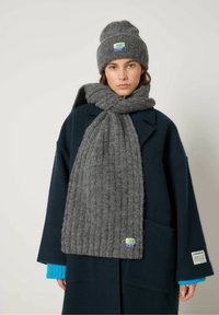 Grå strikket hue og tørklæde sæt, lavet af struktureret stof med ribbet mønster. Tørklædet lægger sig over en marineblå frakke, der har blå kantdetaljer ved ærmerne.