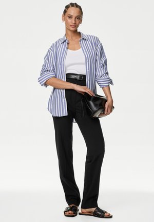 Mujer con camisa abotonada a rayas azules y blancas sobre camiseta sin mangas blanca, pantalones negros con cinturón, sosteniendo un bolso negro y llevando sandalias negras.