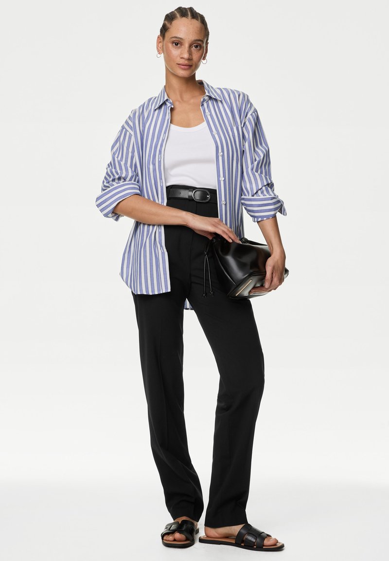 Kobieta w niebiesko-białej koszuli w paski z guzikami, na białym topie, czarne spodnie z paskiem, trzyma czarną torebkę i ma na nogach czarne sandały na płaskiej podeszwie.