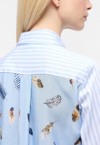 Light blue shirt with a striped collar. Back features a feather print on soft fabric, blending artistic patterns with a relaxed design.