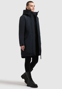Marineblå isoleret parka med aftagelig hætte, featuring sidelommer, en lynlås med en klap, og et knælangt design. Sorte snørebåndsstøvler.