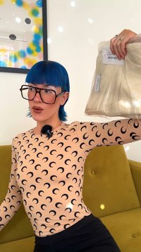 Ročna majica dolgih rokavov v breskovi barvi s črnim polaščnim vzorcem, ki drži bež paket tkanine. Ozadje prikazuje zeleno kavč in umetnine.