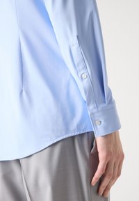 Camisa de manga larga azul claro con puños abotonados. Tejido suave, corte ajustado y un dobladillo ligeramente curvado visible junto a unos pantalones grises.