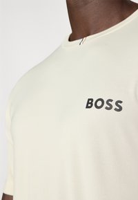 Svetlo bež majica s kratkimi rokavi, izdelana iz teksturirane tkanine, z black "BOSS" logotipom na prsih in majhnim črtastim detajlom na izrezu.