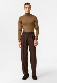 Bruine turtleneck trui gecombineerd met hooggesneden, plooibare bruine broek. Model draagt donkerbruine schoenen. Eenvoudig, aansluitend ontwerp.