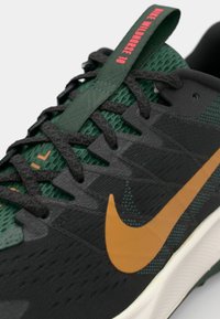 Chaussure de sport noire et verte avec une tige texturée, présentant un grand swoosh orange de Nike et des lacets proéminents. Inclut un onglet vert sur la languette.