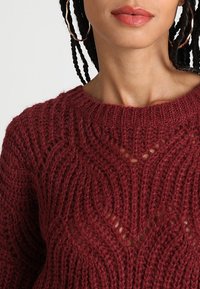 Sweter w kolorze wina o teksturowanym, dzierganym wzorze, z okrągłym dekoltem i otwartymi oczkami tworzącymi falisty wzór.