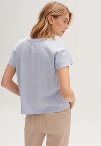 Blusa azzurro chiaro a maniche corte con una sottile trama, scollatura rotonda e dettaglio a pieghe sul retro, abbinata a pantaloni beige.