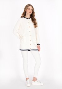 Cardigan crem cu accente bleumarin și nasturi aurii. Prezintă două buzunare frontale, mâneci lungi și o croială lejeră, asortat cu colanți albi.