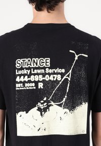 Black t-shirt featuring a white graphic print of a lawn mower and text: "STANCE Lucky Lawn Service" with phone number and establishment year.