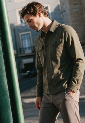 Homme aux cheveux bruns portant une veste verte boutonnée et un pantalon beige se tient à l'extérieur, tête inclinée vers le bas et mains dans les poches.