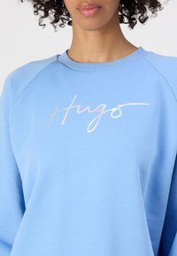 Felpa azzurra chiara caratterizzata da una texture morbida, maniche raglan e un logo decorativo lucido "Hugo" sul petto.