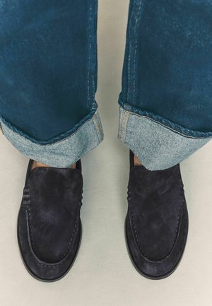 Mocassini navy in suede con dettagli cuciti, abbinati a jeans scuri in denim risvoltati che mostrano una leggera fodera interna. Design minimalista, suola piatta.