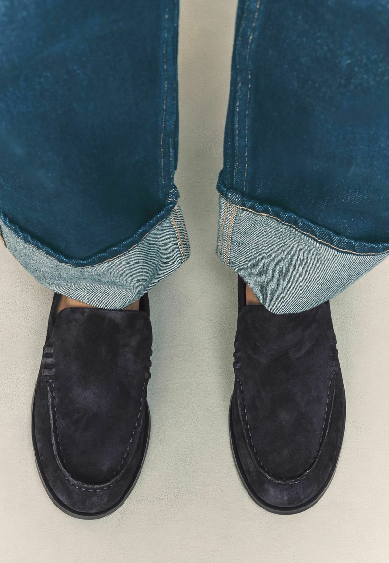 Navy suede loafers with stitched accents, paired with cuffed dark denim jeans showing a light inner lining. Minimalist design, flat soles.