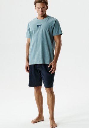 Młody mężczyzna stoi boso, ubrany w morski turkusowy t-shirt z grafiką palmy oraz czarne szorty na białym tle.