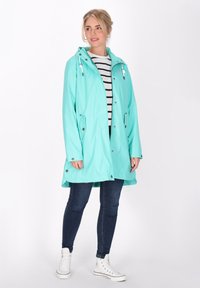 Mentazöld vízálló dzseki kapucnival, elöl cipzárral és oldalsó zsebekkel, csíkos póló felett viselve, sötét farmerral és fehér sportcipővel párosítva.