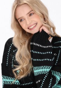 Črna pletena jopica z teal in belimi geometrijskimi vzorci, rebrastimi manšetami in sproščenim fitom. Mehka tekstura, sproščen dizajn.