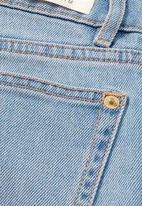 Lichtblauwe denimstof met een zichtbare gouden knop en contrasterende beige stiksel langs de naden en rand van de zakken. Close-up van de textuur.