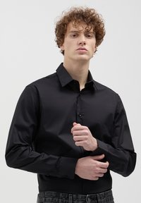 Giovane uomo con capelli ricci che indossa una camicia nera attillata con bottoni, mentre sistema la manica su uno sfondo grigio chiaro uniforme.