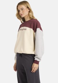 Prugasta dukserica u krem, burgundiji i sivoj boji; ima duge rukave, rebraste manžete i vezeni logo na grudima.