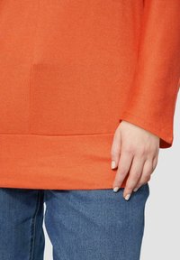 Oranje gebreide trui met een ronde hals en lange mouwen, met een rechte zoom. De hand van het model rust op blauwe denim jeans.