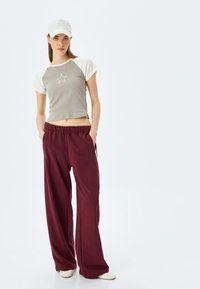 Lys grå og hvid kortærmet T-shirt med et stjerneemblem, parret med løse bordeaux-farvede sweatpants og en hvid cap. Afslappet pasform og casual stil.