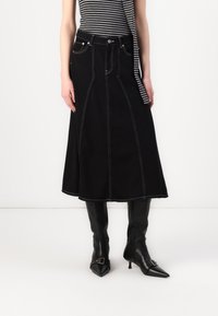 Svart denimskjørt med hvite sømmer, A-linje-design, foran lommer, brukt med knelengde, svarte spisse støvler og en stripete topp.