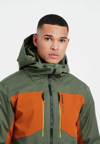 Wasserabweisende Jacke in Olivgrün und Orange. Sie verfügt über einen Reißverschluss vorne, eine verstellbare Kapuze, zwei vordere Reißverschlusstaschen und kontrastierende Reißverschlussakzente.