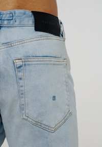 Lyseblå denimjeans med en baklomme og et svart lærmerke med preget tekst. Materialet har en glatt, subtil tekstur.