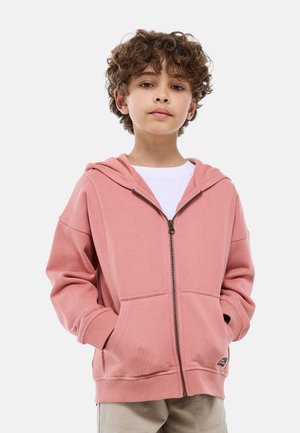 Garçon aux cheveux bouclés portant un sweat à capuche rose zippé sur un t-shirt blanc, se tenant les mains dans les poches devant un fond uni.