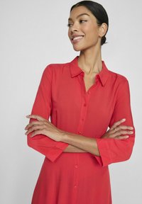 Raudona marškinėlių su apykakle, ilgomis rankovėmis ir lygiu paviršiumi. Turi laisvą siluetą ir nedidelį blizgesį.