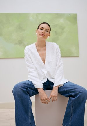 Žena so zastrčenými vlasmi, oblečená v textúrovanom bielom saku a širokých modrých džínsach, sedí na bielom kockovom podstavci pred zeleným abstraktným umením.