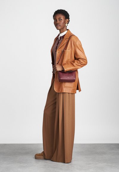 Blazer de couro castanho sobre um suéter vinho escuro, combinado com calças largas castanhas. Uma pequena mala burgundy e sapatos de plataforma completam o visual.