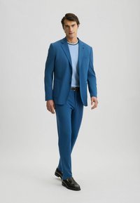 Blauw pak met een slim fit, voorzien van een notch lapel en twee voorzakken. Gemaakt met een lichtblauw overhemd en zwarte instappers. Soepele textuur.