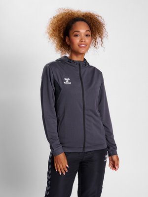 Vrouw met krullend haar, gekleed in een donkergrijze hoodie met rits en zwarte sportbroek met zijstrepen in chevronpatroon, staand tegen een effen achtergrond.