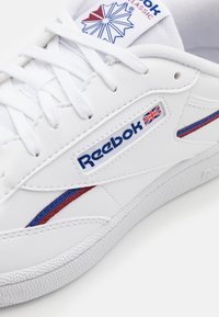 Tênis brancos de couro com detalhes em azul e vermelho, logotipo da Reebok com a bandeira do Reino Unido, sola texturizada e design de cadarço.