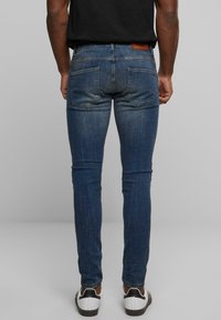 Jeans de mezclilla azul oscuro de corte ajustado con un lavado desgastado, dos bolsillos traseros y un parche de cuero marrón en la cinturilla. Combinados con zapatillas blancas.