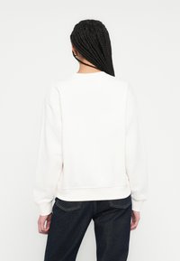 Biela mikina s okrúhlym výstrihom, rebrovanými manžetami a lemom, s hladkou textúrou. Nosí sa s tmavými džínami, zobrazená zozadu.