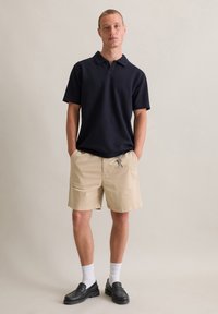 Navy polo shirt in textured fabric, khaki shorts, black leather loafers, and white socks. Pockets visible on shorts, relaxed fit overall.