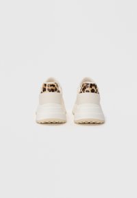 Ett par krämfärgade sneakers med tjocka sulor och detaljer i leopardtryck på hälen, visade bakifrån mot en vit bakgrund.