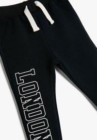 Zwarte joggingbroek met crèmekleurige trekkoorden, met "LONDEN" in vet witte letters op het linkerpijp. Gemaakt van zachte, ademende stof.