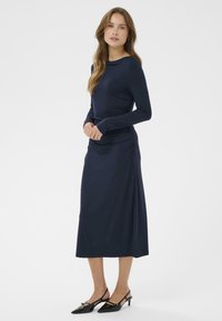 Langärmliges, marineblaues Kleid mit rundem Ausschnitt, gerafftem Seitendetail und geradem Rock. Schwarze Slingback-High Heels mit spitzem Zeh.