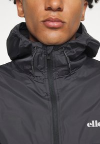 Schwarze Kapuzenjacke mit Reißverschluss vorne, aus leichtem Material gefertigt. Verfügt über eine Kordel am Hals und ein kleines Logo auf der Brust.